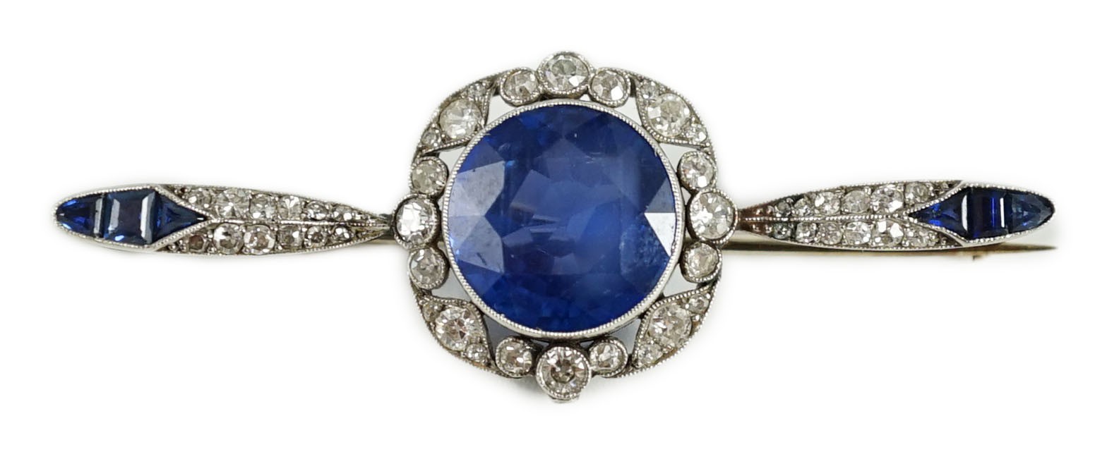 An early 20th century French platinum, sapphire and diamond bar brooch, the central round cut sapphire of good colour, with diamond border, flanked by diamonds and shaped cut sapphires